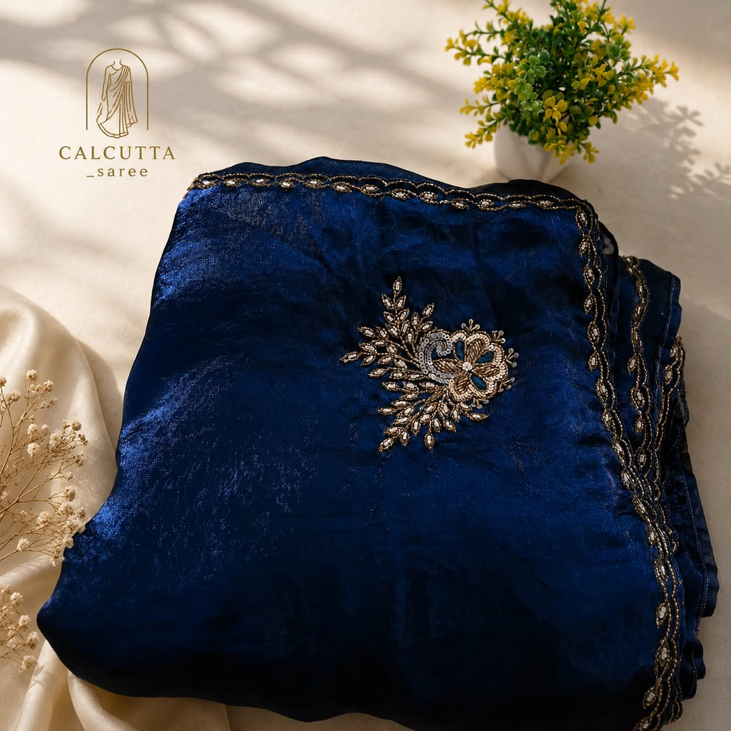 A folded navy blue velvet garment with ornate gold and silver embroidery, displayed on a cream satin fabric with decorative dried flowers and a small potted plant. The brand logo 'CALCUTTA saree' is visible in the top left corner.