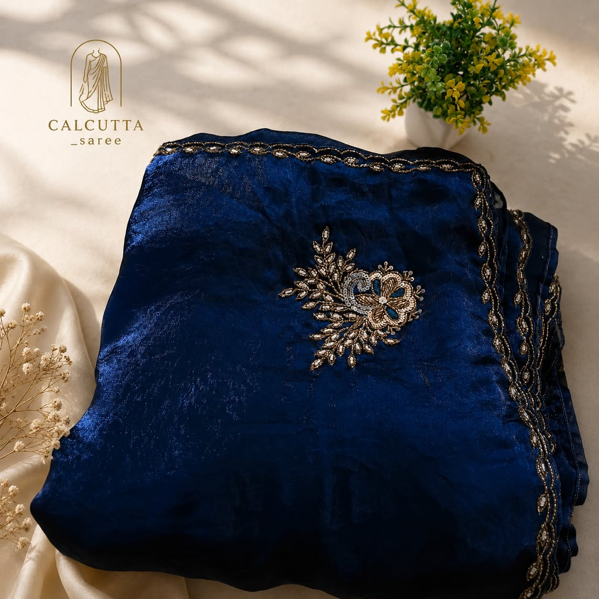 A folded navy blue velvet garment with ornate gold and silver embroidery, displayed on a cream satin fabric with decorative dried flowers and a small potted plant. The brand logo 'CALCUTTA saree' is visible in the top left corner.