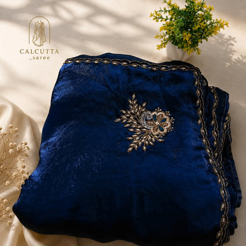 A folded navy blue velvet garment with ornate gold and silver embroidery, displayed on a cream satin fabric with decorative dried flowers and a small potted plant. The brand logo 'CALCUTTA saree' is visible in the top left corner.